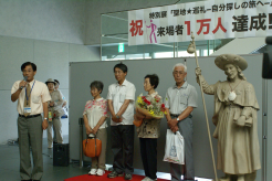 館長からの祝福の言葉を聞く1万人目のお客様とヤコブ像